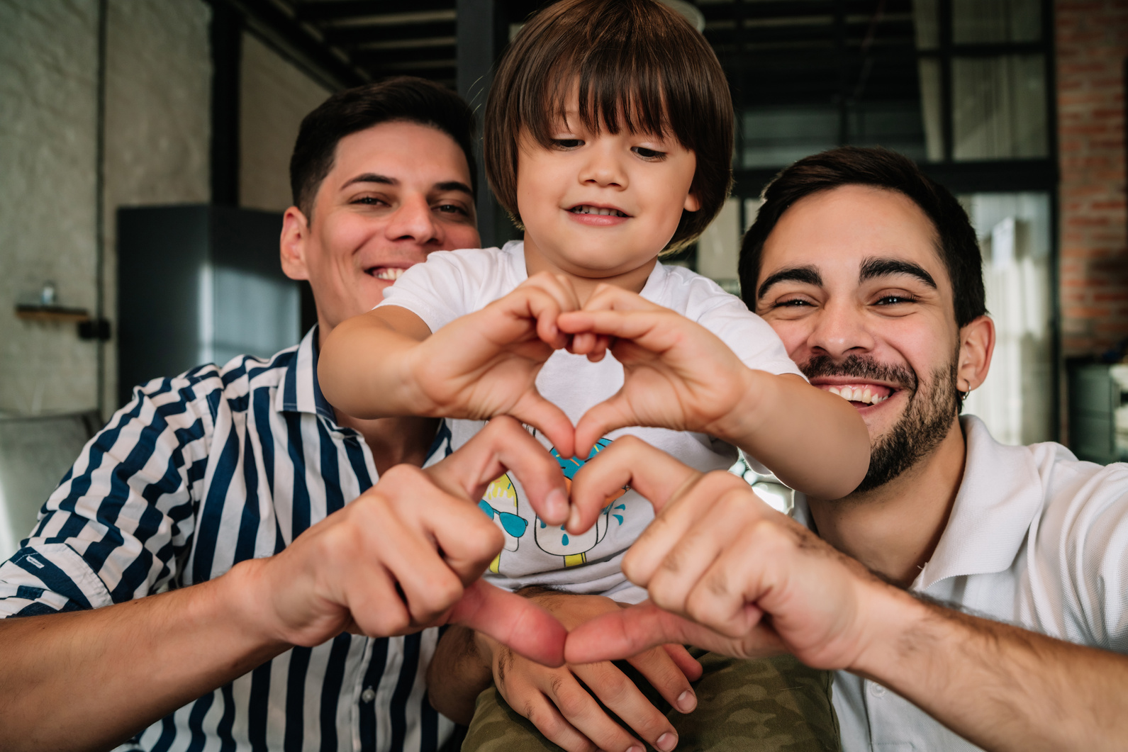 A smiling gay couple poses with their son. They are making heart shapes with their hands.