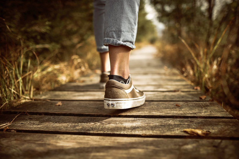 A person is walking on a brown wooden pathway in a nature setting. The image shows only the calves down of the person. They are wearing skate shoes and ankle socks with Capri length jeans.